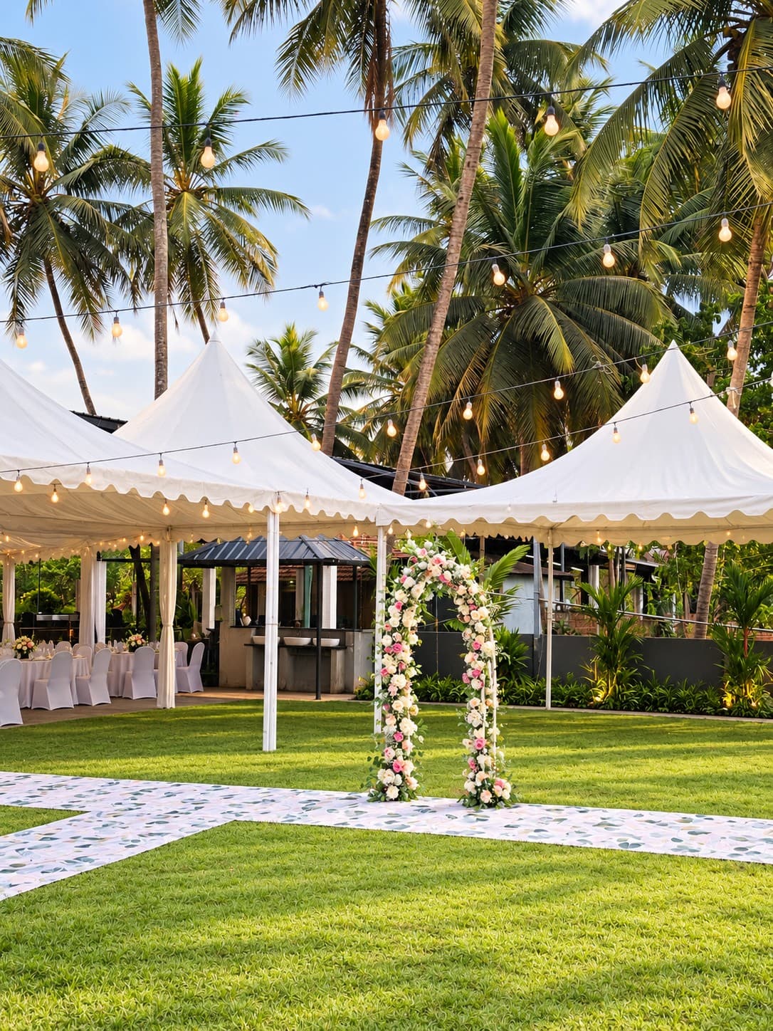The lawn set for a wedding reception: white peaked canopies with tables and covered chairs beneath them, a flower arch of pink and white roses over a printed aisle runner on the grass, and string lights hung through the coconut palms.