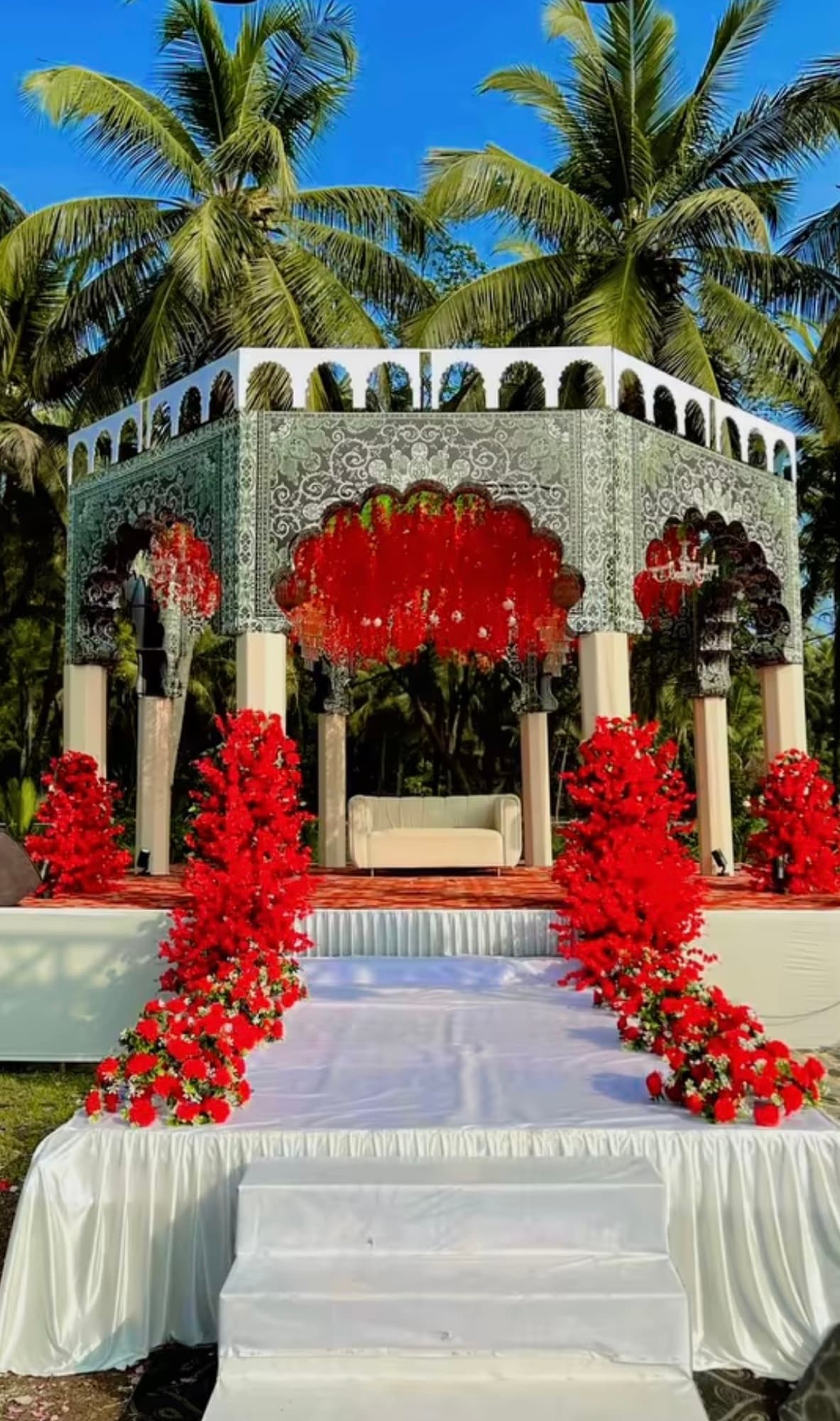A wedding mandap on the grounds: a white and silver filigree pavilion under coconut palms, banked with red roses, a cream sofa on the stage and a white carpet running up the steps to it.