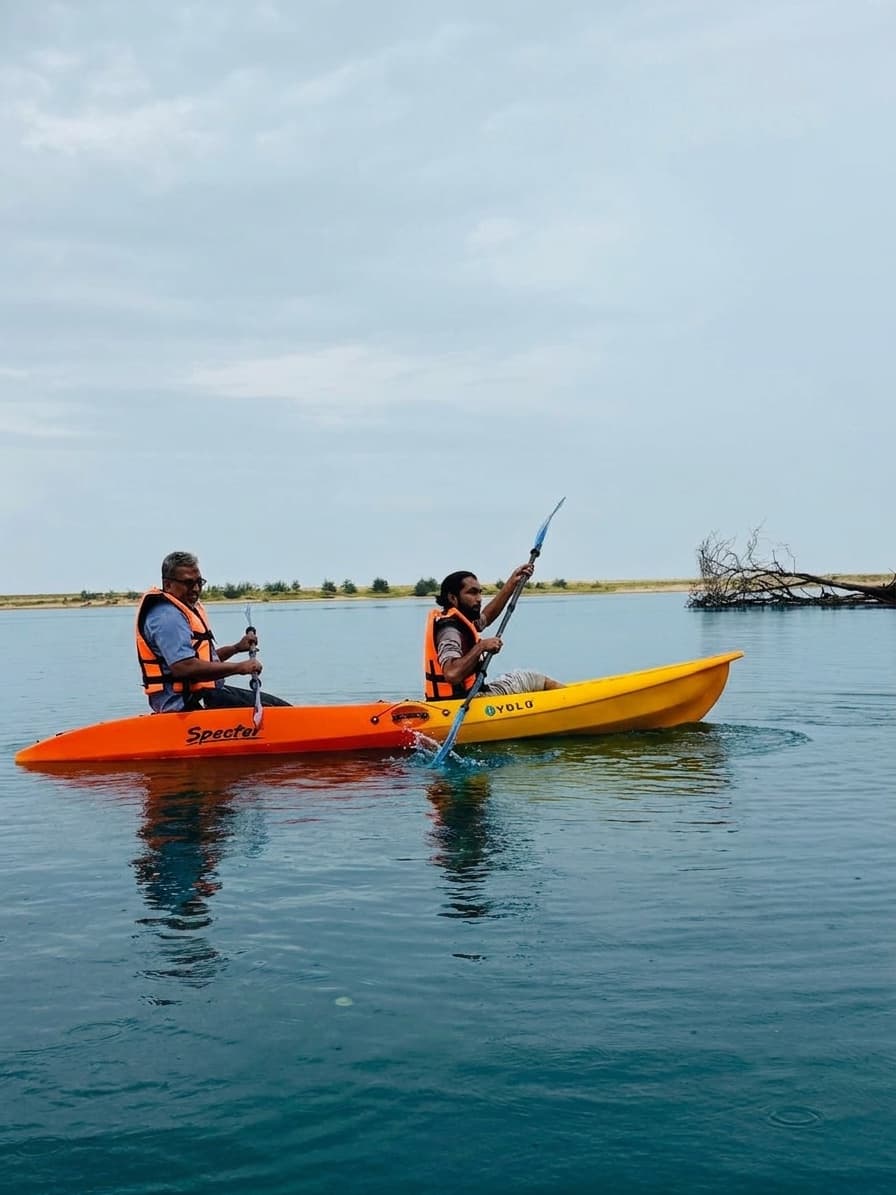 Two paddlers in orange life jackets in an orange and yellow two-seater sit-on-top kayak on the flat blue-green water of the Kumbla estuary, one paddle raised mid-stroke, with a low sandy shoreline and bare driftwood behind them.